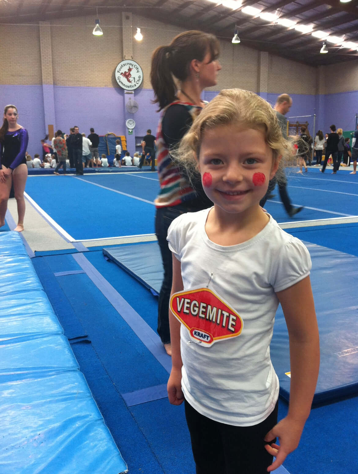 Smiling girl with rosy cheeks at a gymnastics event, wearing a Vegemite shirt, surrounded by gymnasts in the background.